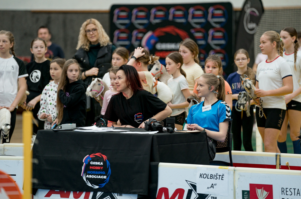 Drukke indoor Hobby Horsing-evenementsscène met twee organisatoren aan een zwarte tafel met het logo van Česká Hobby Horsing Asociace, terwijl meisjes in sportkleding eromheen staan met hun Hobby Horses. Witte pisteafzettingen, het bord Kolbiště „A“ en