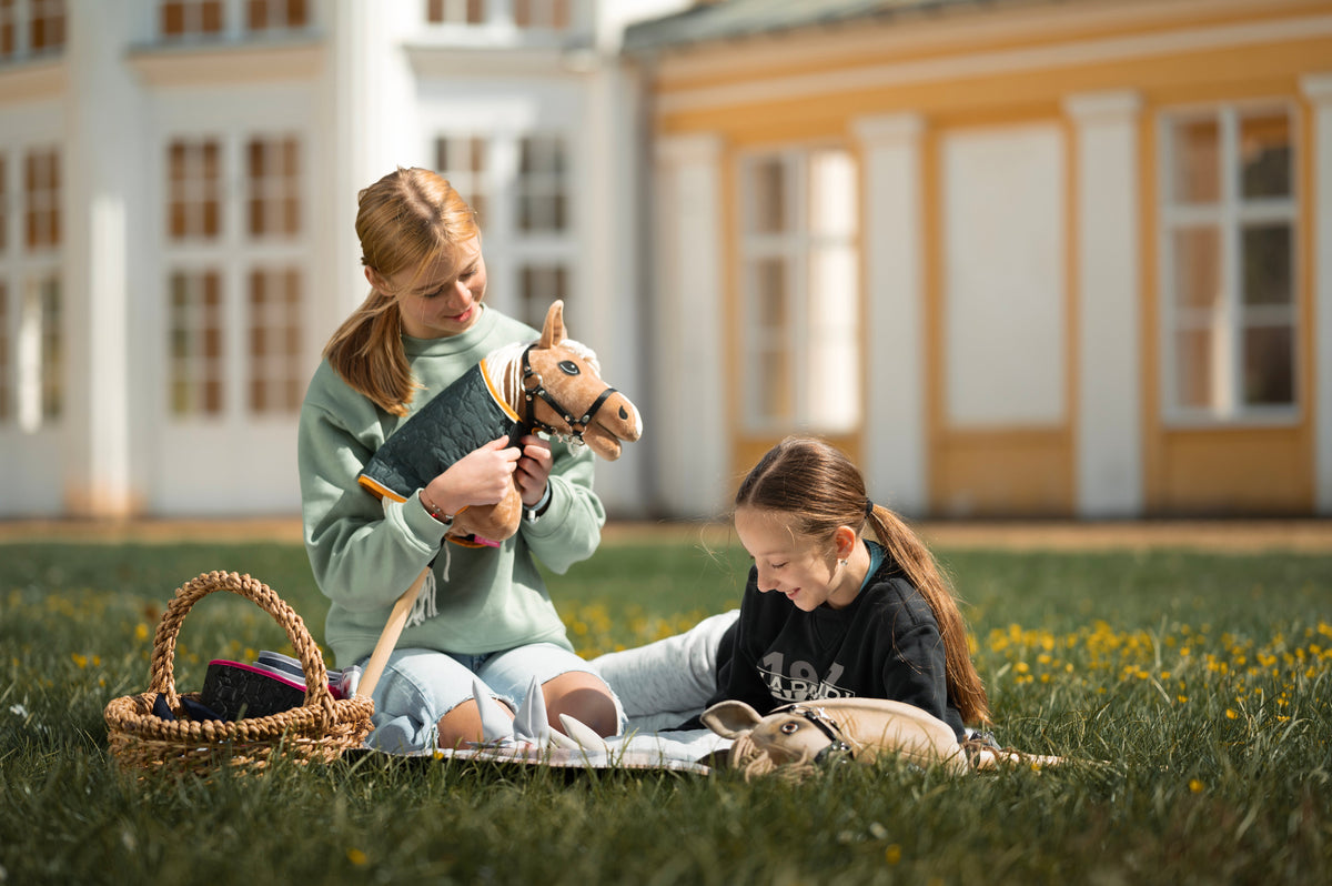 Une fille en sweat vert pâle est dans l’herbe près d’un panier en osier d’accessoires, tenant un Hobby Horse beige à Bride noire, crinière blanche et couverture matelassée, un enfant est visible sur une couverture