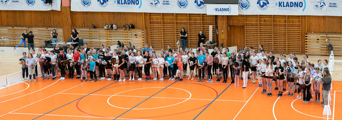 Grand groupe de jeunes participants au Hobby Horsing alignés sur un terrain de sport intérieur orange, en tenue décontractée sportive et tenant divers Hobby Horses, avec espaliers en bois, gradins et banderoles de Kladno à l’arrière-plan