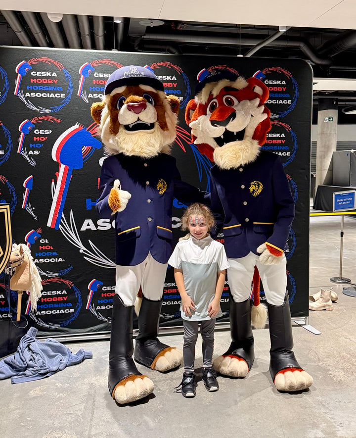 Jeune fille souriante au visage peint entre deux mascottes animales de Prague Playoffs en vestes bleu marine style équitation et casquettes, devant un fond de Česká Hobby Horsing Asociace dans le hall de l’O2 Arena, avec un petit Hobby Horse à côté