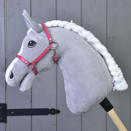 Un Hobby Horse gris en peluche, liste blanche et crinière tressée, porte le licol rose framboise devant un mur en bois gris
