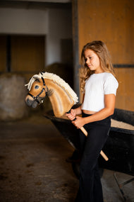 Meisje in een wit topje staat in een stal naast een kruiwagen en houdt Hobby Horse Colby in zwarte halster vast