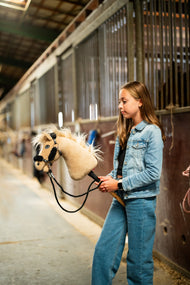 Meisje in spijkerjack en jeans houdt een crèmekleurige Hobby Horse met zwart halster met bont aan de zwarte teugels in de stalgang