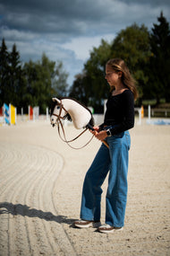 Fille sur une carrière en sable avec obstacles colorés tenant un Hobby Horse crème en bride complète marron avec muserolle