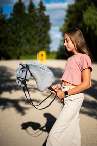 Meisje in een roze shirt loopt over een zandbak met Hobby Horse Grace in zwart hoofdstel met teugels