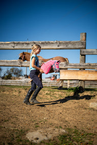 Lachend meisje in rijlaarzen loopt met Hobby Horse Pinkie, hier met turquoise halster, over de paddock langs een vos bij het hek