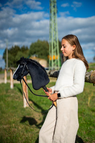 Meisje in een witte pluizige trui in een zonnige wei houdt Hobby Horse Loki vast in een zwart hoofdstel met teugels