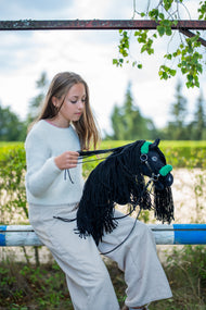 Meisje in witte trui zit op een balk en pakt de teugels op van de zwarte Hobby Horse in een groen halster met bont