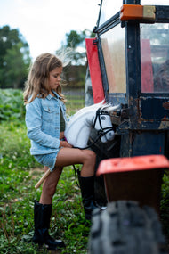 Fille en veste en jean et bottes noires près d’un vieux tracteur, tenant le Hobby Horse Nexus en bride noire avec rênes