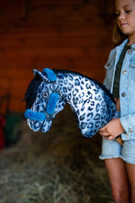 Fille en veste en jean dans une écurie tenant un Hobby Horse à motif léopard bleu et blanc avec le licol bleu pétrole en fourrure