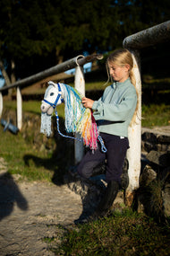 Een meisje in mintgroene sweater bij het witte hek van de rijbak houdt een witte Hobby Horse met regenboogmanen aan het blauw gestreepte halstertouw