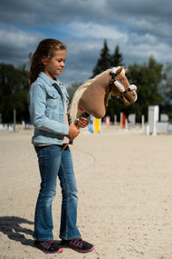Meisje in spijkerjasje op een zandrijbaan met hindernissen met een lichtbruine Hobby Horse in de halster met crèmekleurig bont