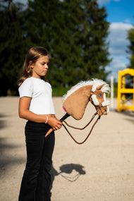 Girl in a white top on a sand arena holding Hobby Horse Star, here in a brown halter with cream fur and brown rope reins
