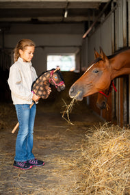 Fille dans une écurie tenant un Hobby Horse brun moucheté au licol bordeaux avec fourrure, à côté d’un cheval bai qui mange du foin