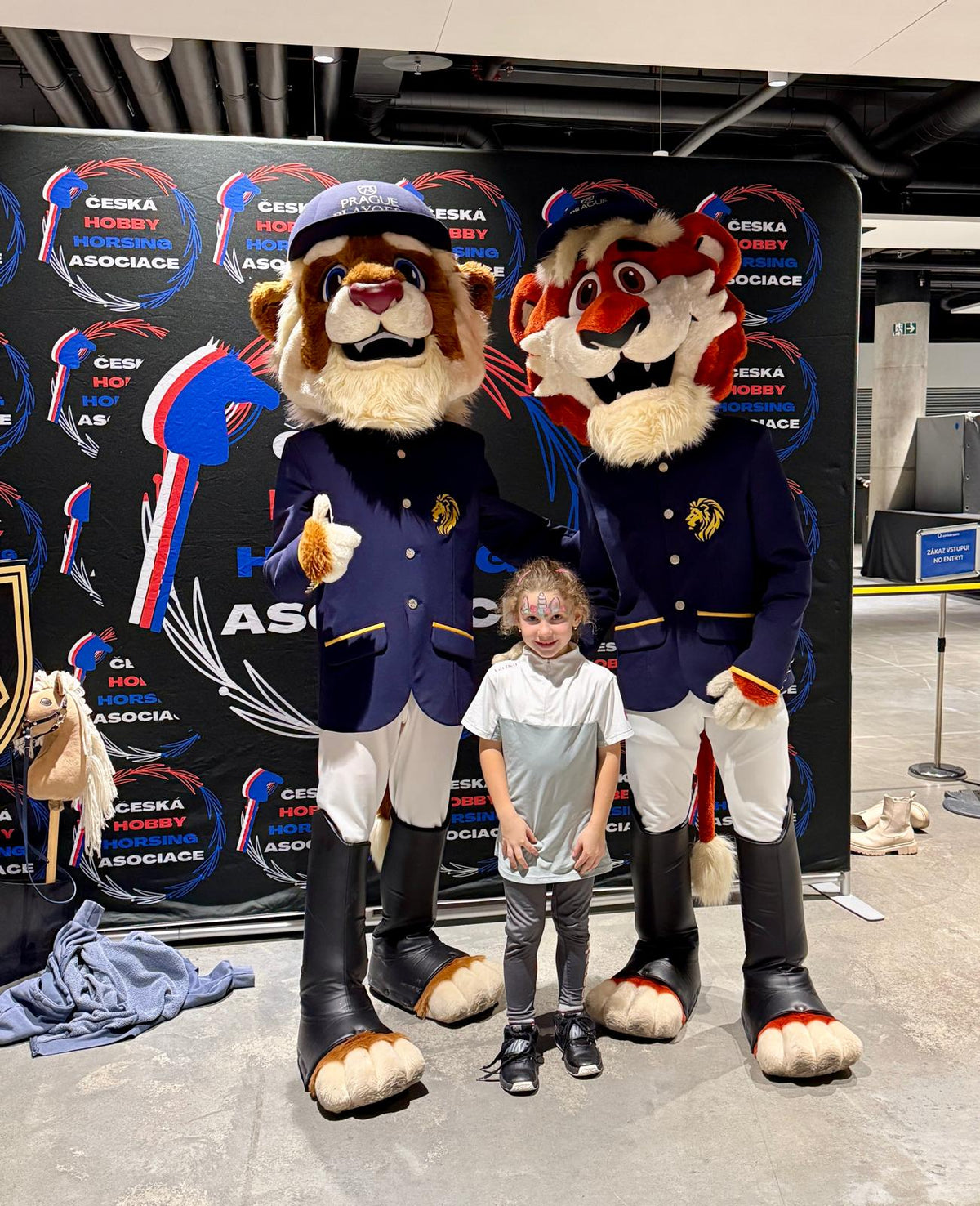 Jeune fille souriante au visage peint entre deux mascottes animales de Prague Playoffs en vestes bleu marine style équitation et casquettes, devant un fond de Česká Hobby Horsing Asociace dans le hall de l’O2 Arena, avec un petit Hobby Horse à côté