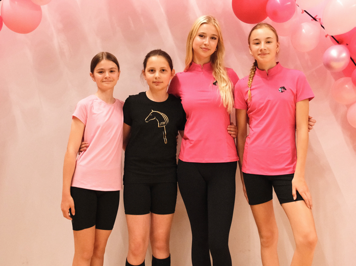 Quatre jeunes debout bras dessus bras dessous devant un fond rose pâle décoré de ballons roses, portant des hauts sportifs roses et noirs avec shorts ou leggings noirs et des détails de logo Hobby Horse