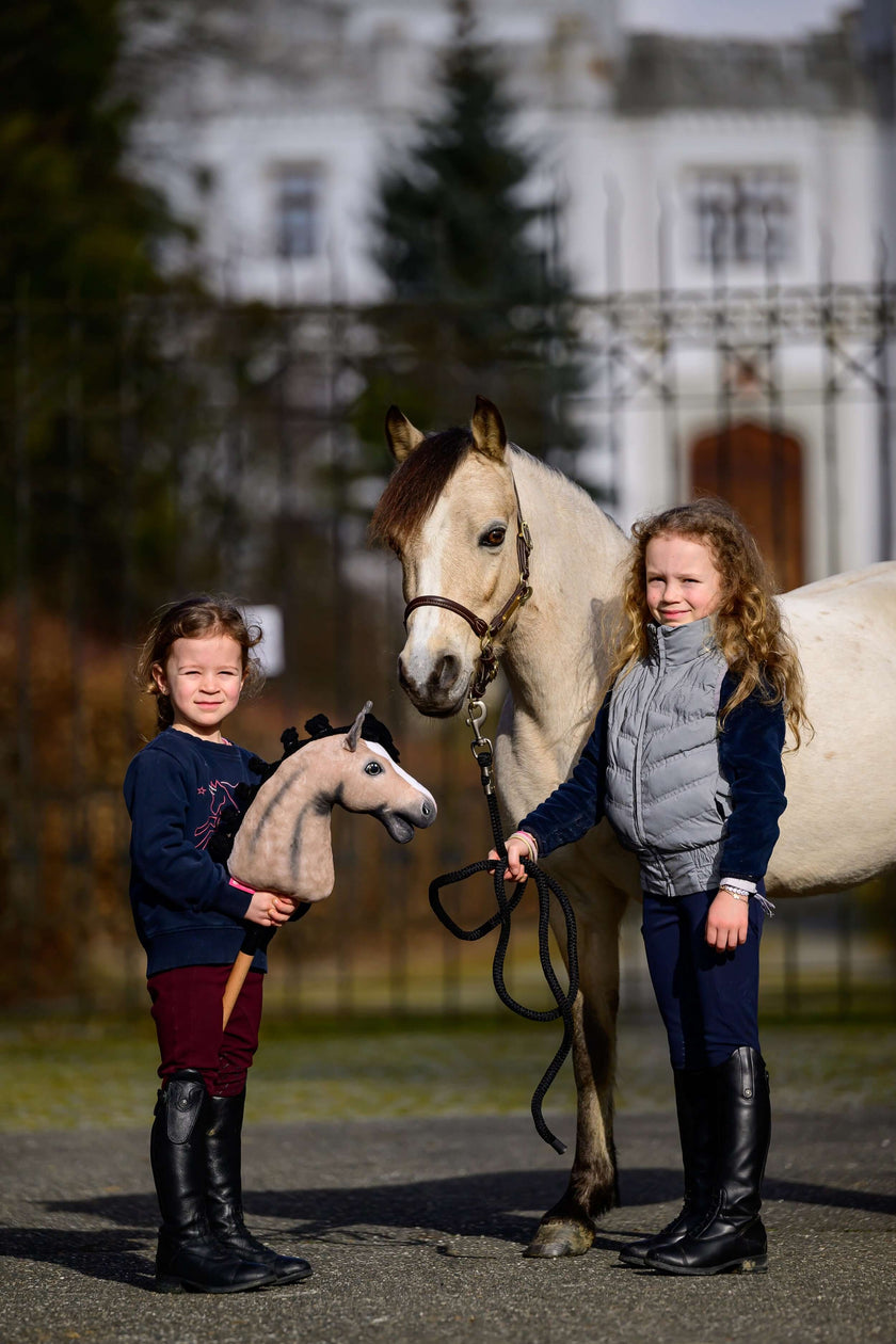Two girls in riding boots, one with a beige Hobby Horse, the other holding a cream pony on a lead rope