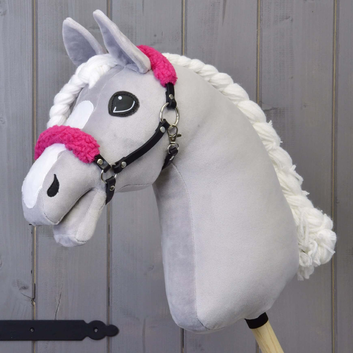 Een grijze pluche Hobby Horse met witte bles en gevlochten manen draagt de zwarte halster met krullend felroze bont
