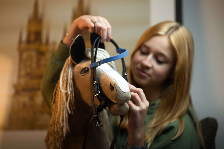 Blonde girl fitting a bridle with a blue noseband on her light brown Hobby Horse with a cream yarn mane