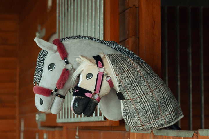 Grey Hobby Horse in a burgundy fur halter beside a cream one in a pink halter and grey check blanket in a stable