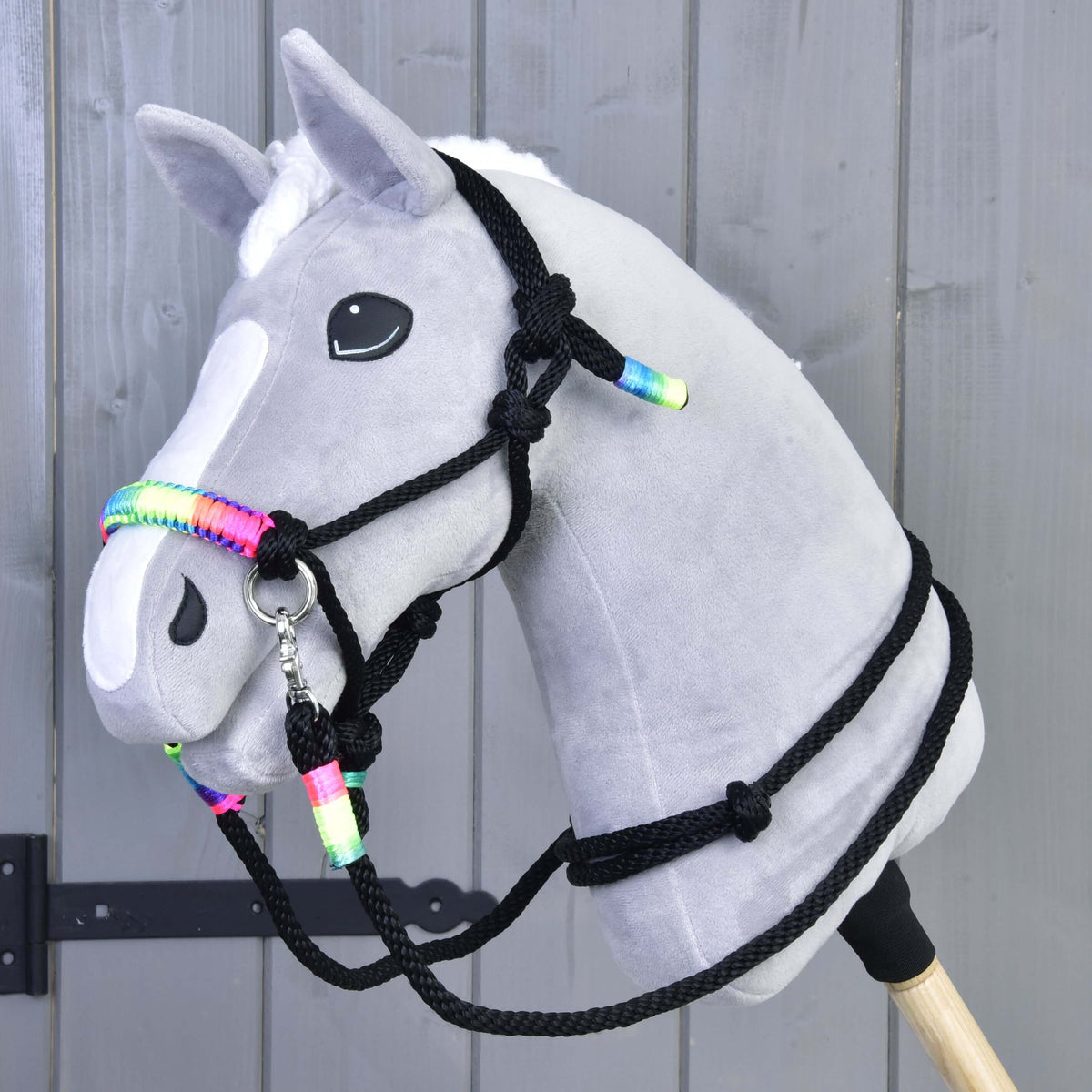 Hobby Horse gris à liste blanche avec licol et rênes en corde noire à gaines arc-en-ciel devant un mur en bois gris