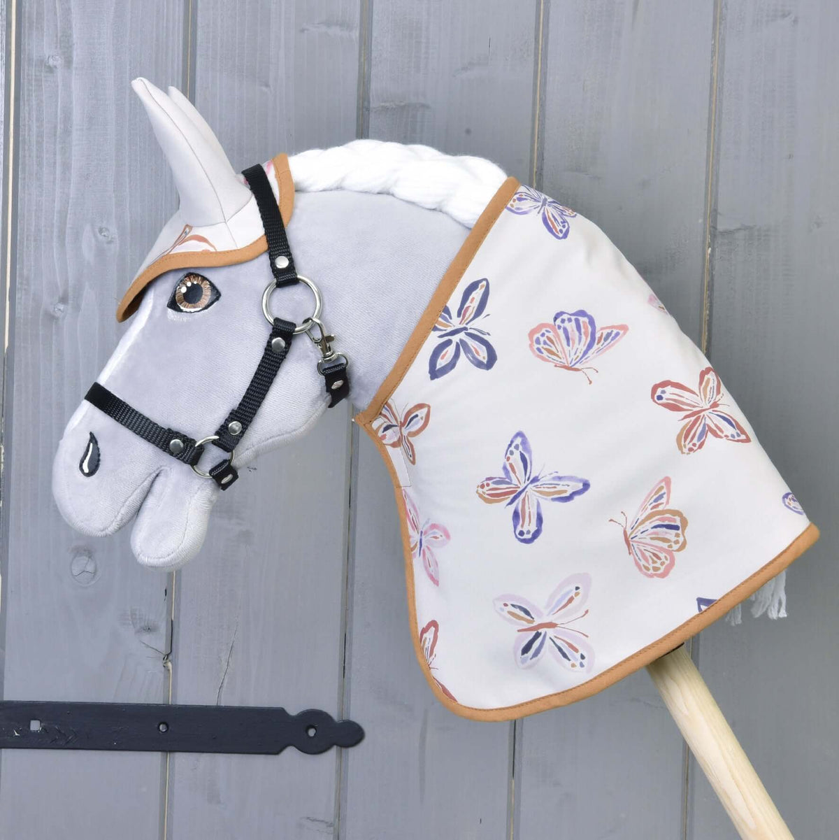 A grey plush Hobby Horse wears the white butterfly blanket with a matching ear bonnet and a black halter by a grey wall