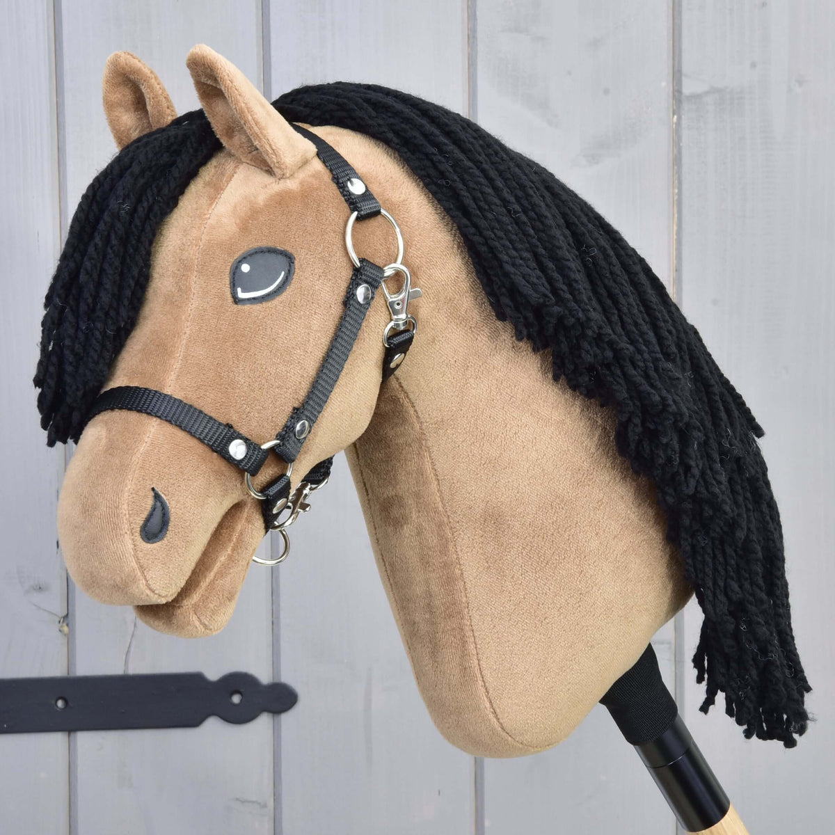 LarDen Hobby Horse Samantha met zwart halster, zandkleurig pluche hoofd en lange zwarte manen van garen voor een grijze houten wand