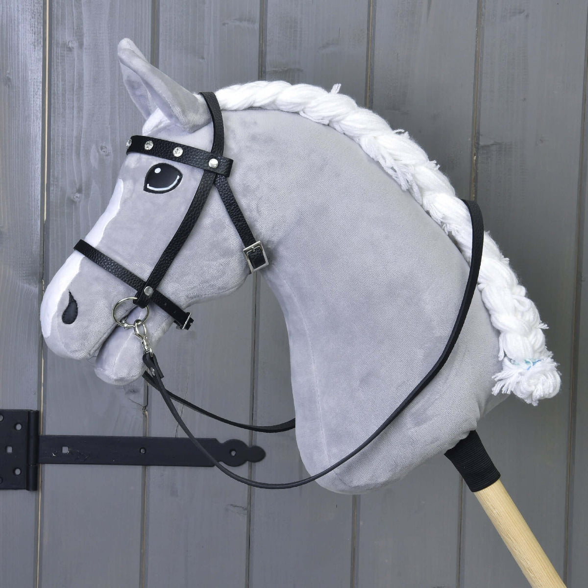 Side view of a grey plush Hobby Horse with a braided mane in the black bridle with rhinestones and reins