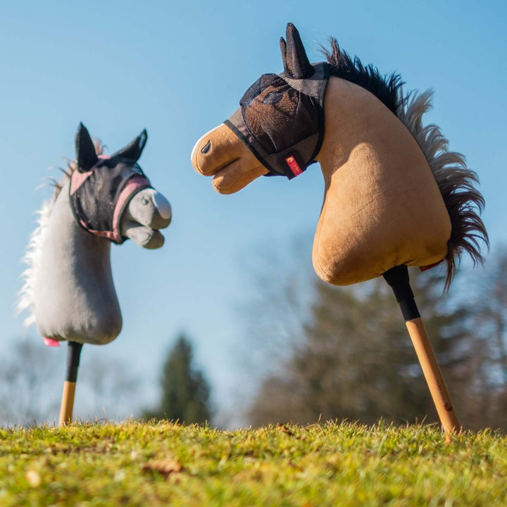 Grey and light brown Hobby Horses wearing black mesh fly masks with ears on a grassy hill under a blue sky