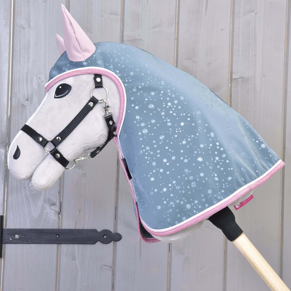 Lichtgrijze Hobby Horse met zwarte halster draagt de grijsblauwe regenjas met capuchon, roze oren en roze bies