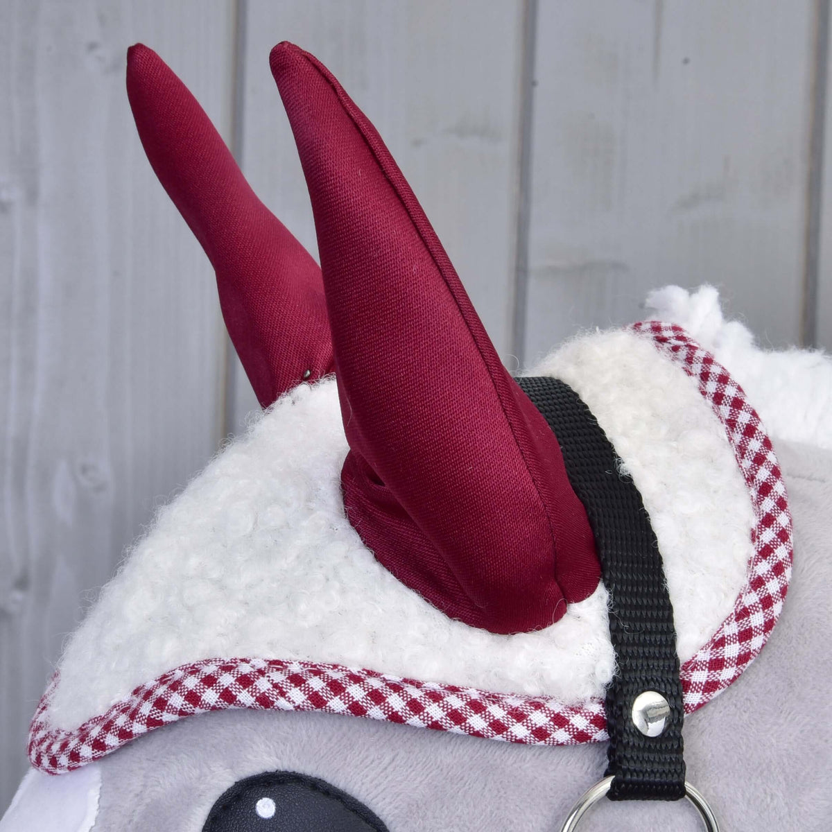 Bordeauxrode oren en rood geruite rand van het oornetje van witte teddy op een grijze Hobby Horse met zwart halster