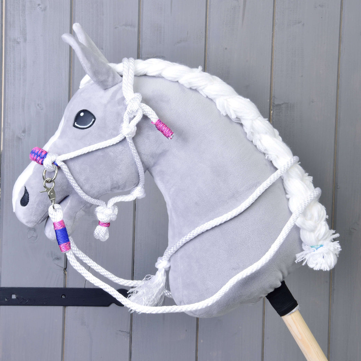 Side view of a grey Hobby Horse in the white rope halter and reins with pink and blue wraps, grey wooden wall