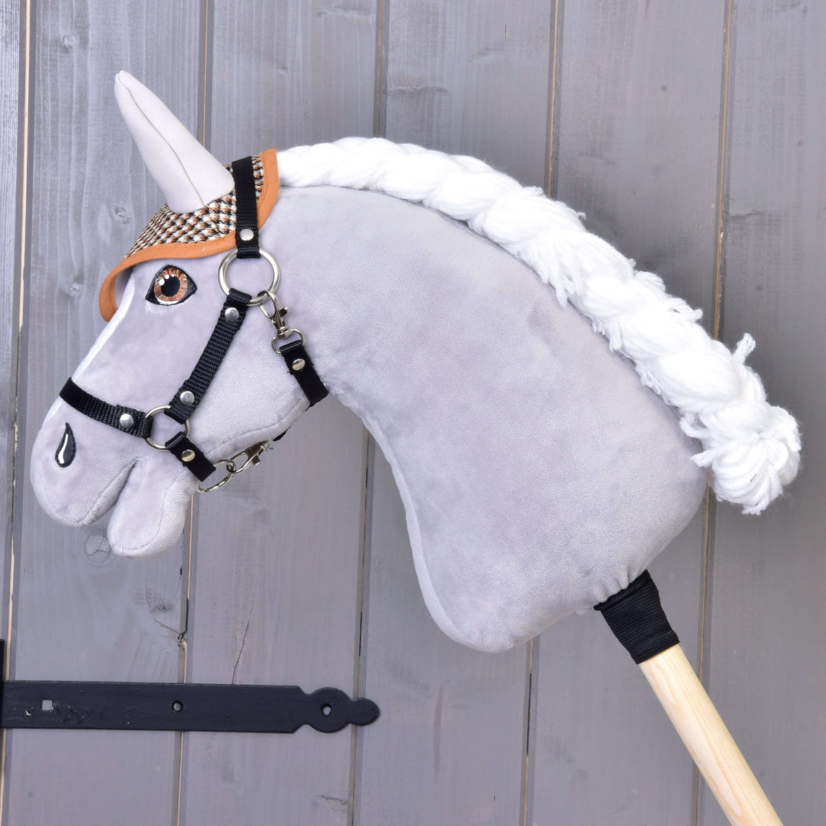 Een grijze pluche Hobby Horse met witte gevlochten manen en zwarte halster draagt het bruine pied-de-poule oornetje