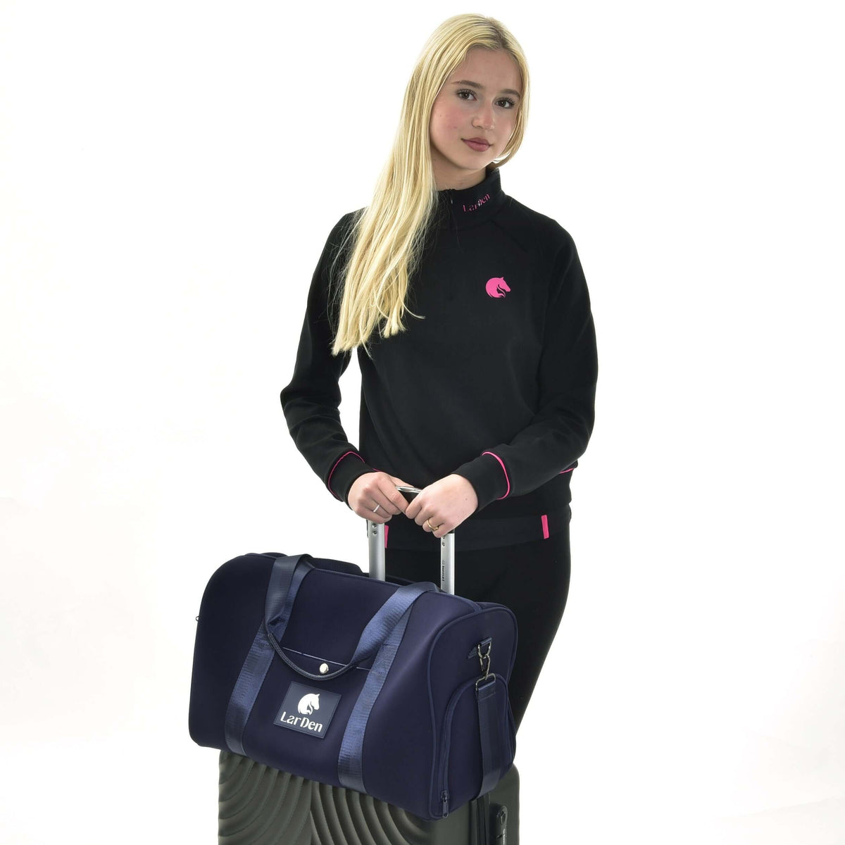 Jeune fille blonde en sweat-shirt noir à détails roses tenant une valise surmontée du sac de sport Hobby Horse bleu marine