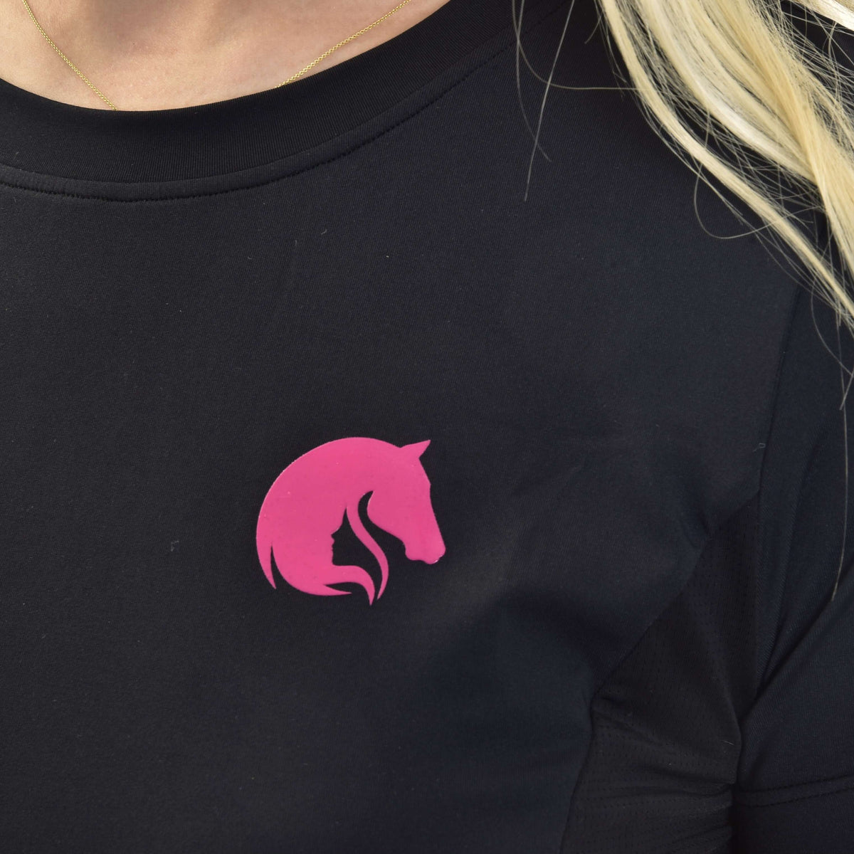 Close-up van het roze paardenhoofdlogo op een zwart sportshirt met ronde hals, gedragen met een dun gouden kettinkje