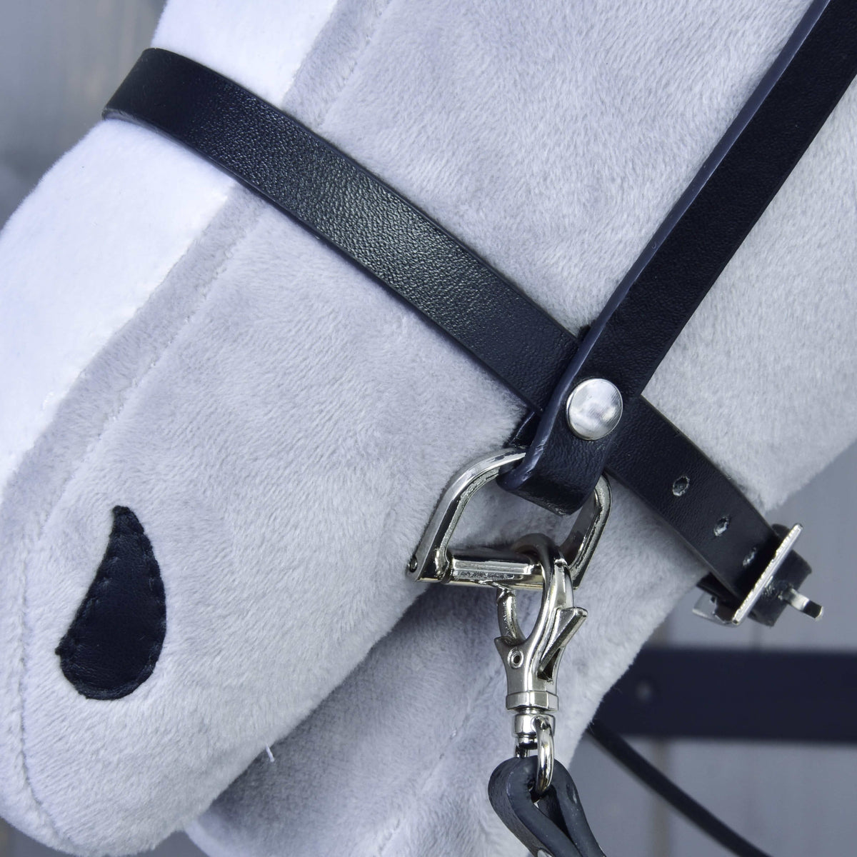 Detail van de zwarte neusriem van kunstleer met zilveren D-ring en karabijnhaak van de teugels op de neus van een lichtgrijze Hobby Horse