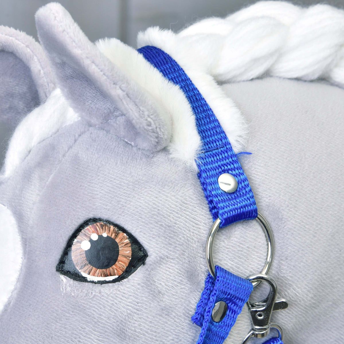 Close-up van het blauwe kopstuk met wit bont achter het oor van een grijze Hobby Horse, met zilveren ring en musketon