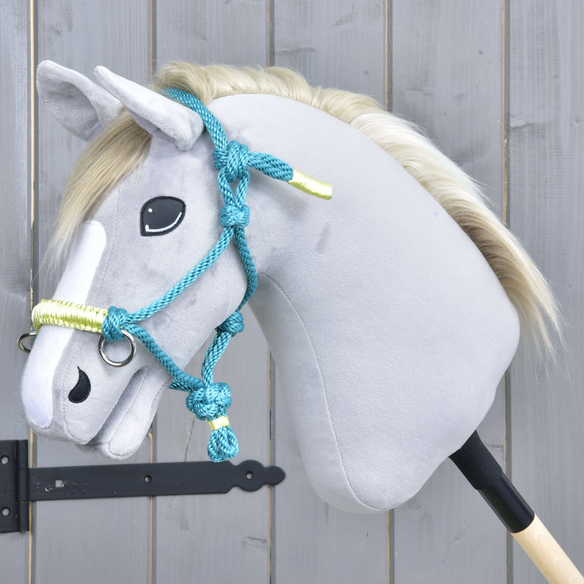 Lichtgrijze Hobby Horse met blonde manen in de turquoise touwhalster met lichtgele neusriem voor een grijze deur