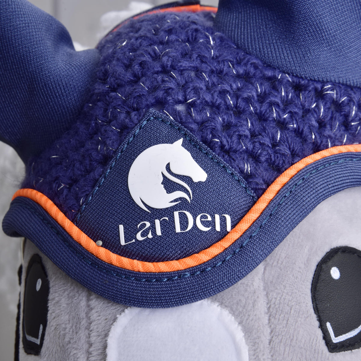 Gros plan sur le logo blanc LarDen à tête de cheval du bonnet d’oreilles au crochet bleu marine, bordé de passepoil orange