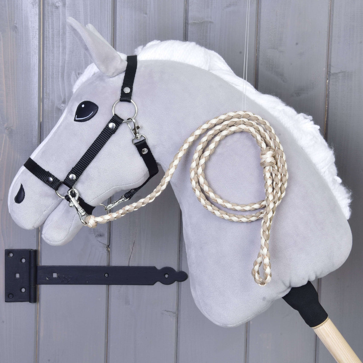Cream and beige braided lead rope clipped to the black halter of a grey Hobby Horse by a grey wooden wall