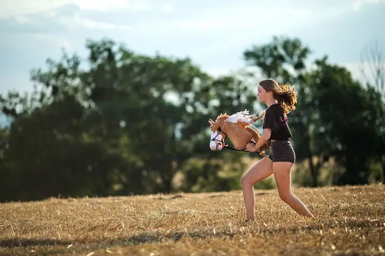 Girl galloping across a dry stubble field with a light brown Hobby Horse with a white mane