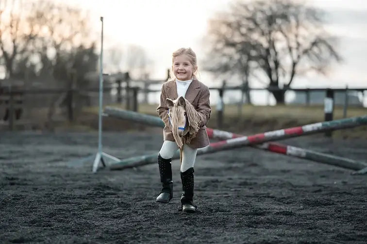 Little girl in a tweed jacket and riding boots playing with a cream Hobby Horse in a sand arena with poles