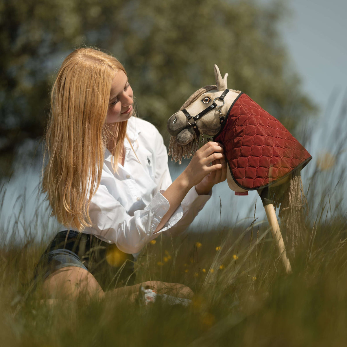 Blonde girl sitting in tall grass with a dun Hobby Horse wearing a black halter and a red quilted blanket