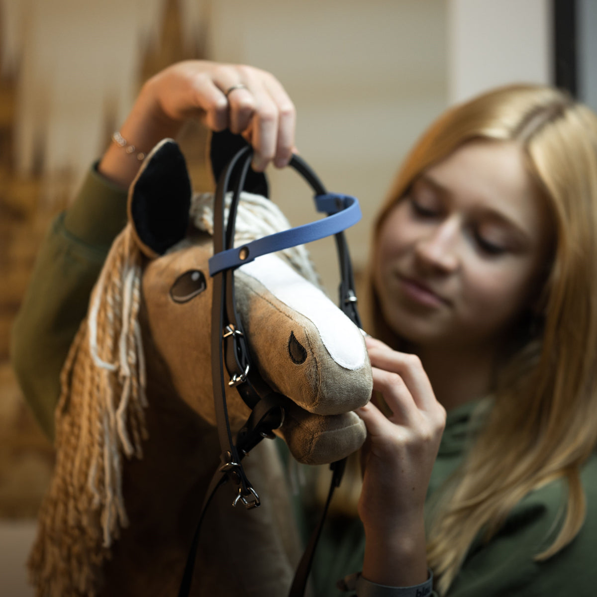 Fille blonde mettant une bride marron foncé à frontal bleu à un Hobby Horse brun clair à crinière blonde en fil