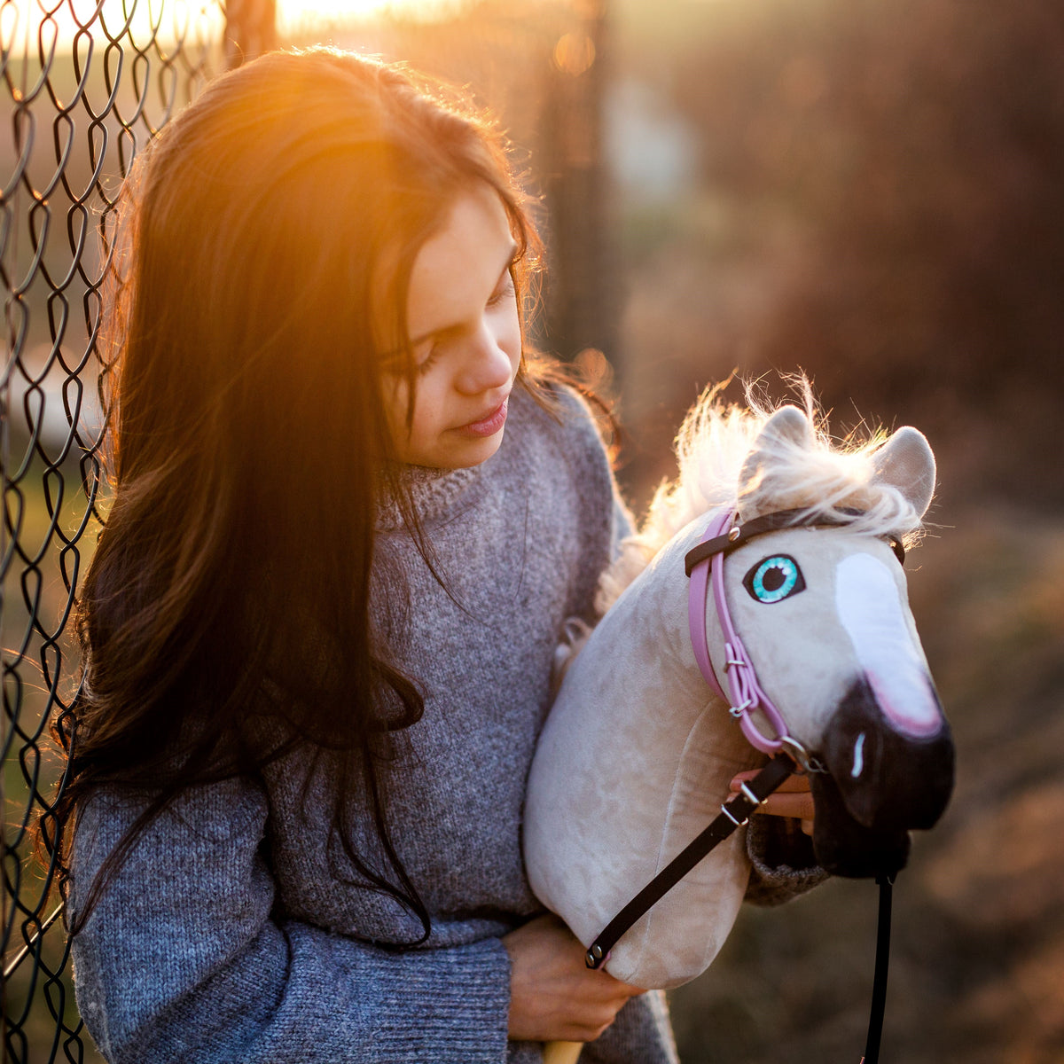 Fille en pull gris tricoté avec un Hobby Horse blanc à la bride rose près d’une clôture dans la lumière dorée du soir