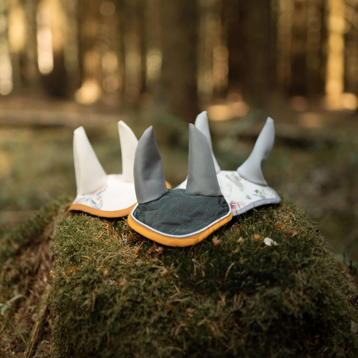Trois bonnets d’oreilles pour Hobby Horse sur une souche moussue en forêt : deux à motif floral et un vert foncé matelassé