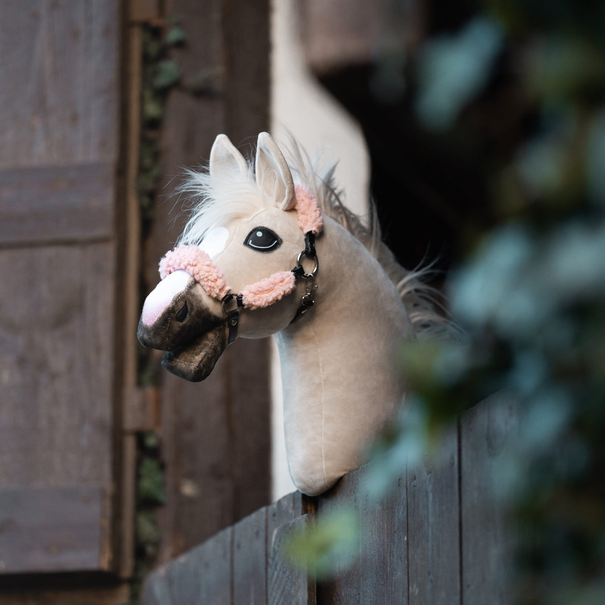 Beige Hobby Horse met zwarte halster met roze bont kijkt over een houten staldeur