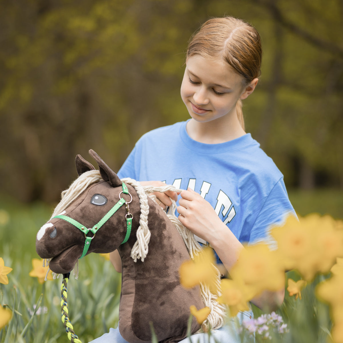 Fille en t-shirt bleu tressant la crinière d’un Hobby Horse brun foncé au licol vert parmi des jonquilles jaunes