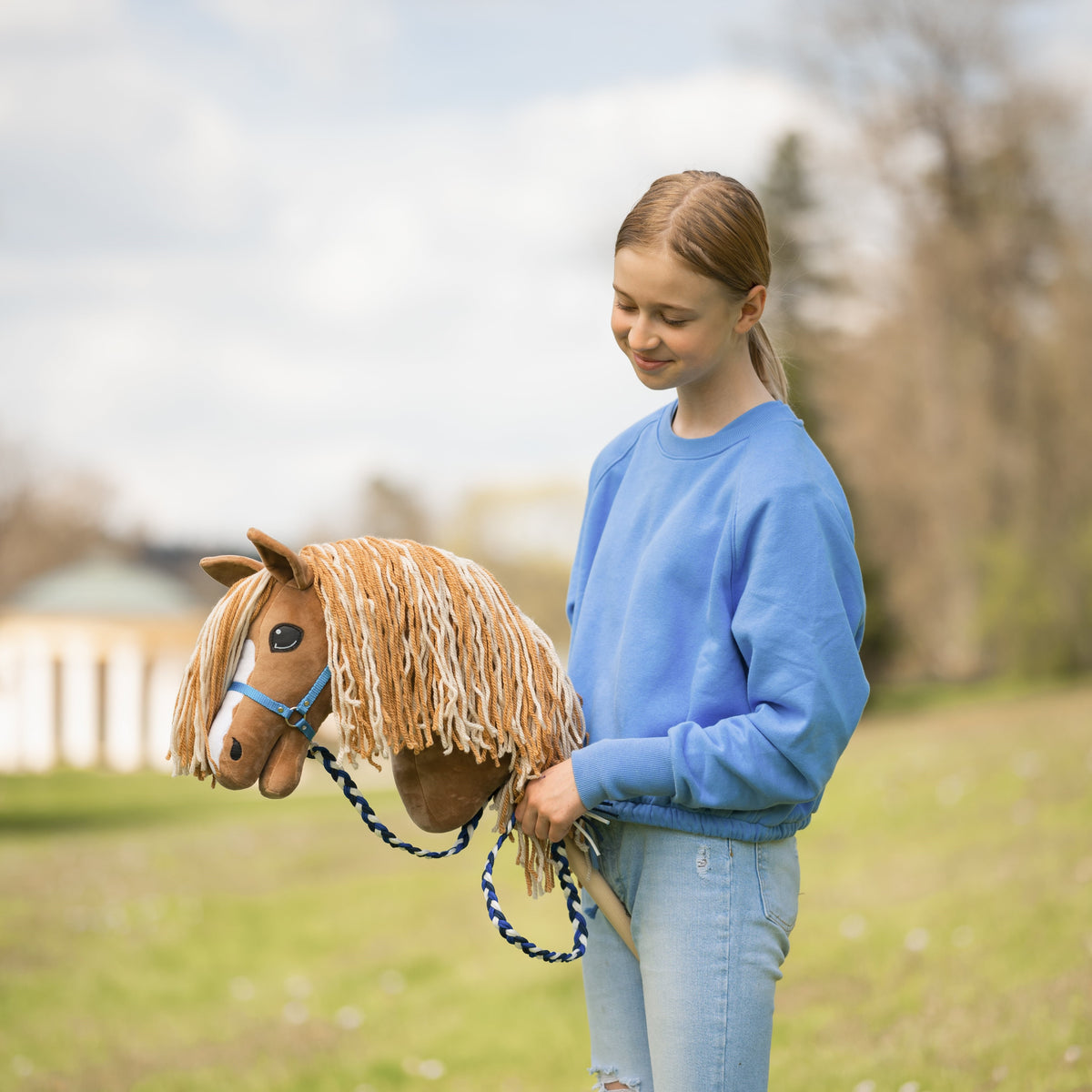 Girl in a blue sweatshirt smiling at a chestnut Hobby Horse with a long blonde mane, blue halter and braided lead rope