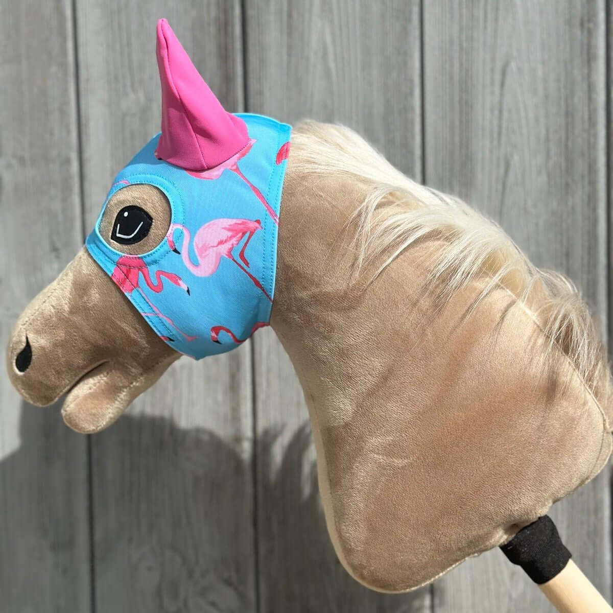 Beige Hobby Horse with a blonde mane wearing a blue mask with a pink flamingo print and a pink ear cover, side view
